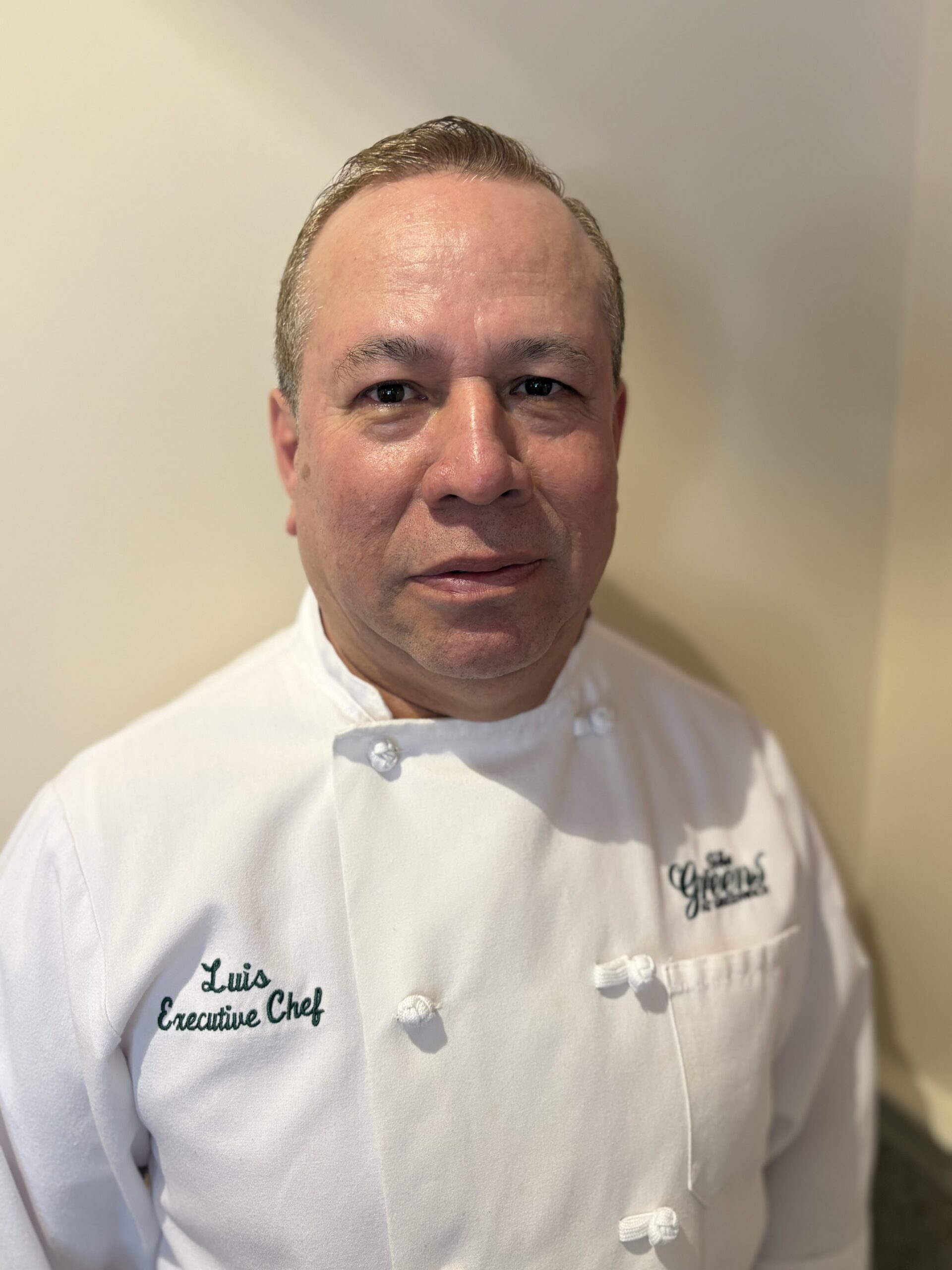 Chef in a white double-breasted jacket with 'Luis Executive Chef' embroidered, standing against a light wall.